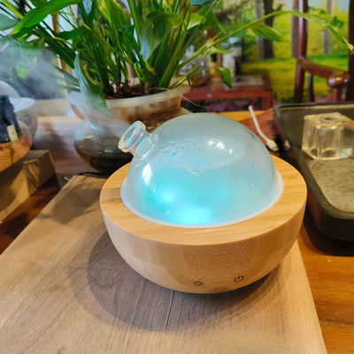 SereneGlow Glass Aromatherapy Diffuser with 200ml glass dome and wooden base emitting blue LED light on wooden table